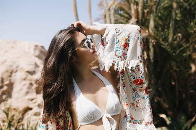 Portrait of amazing slim brunette girl in trendy sunglasses enjoying vacation in tropical country. Graceful young woman in white swimwear has sunbathing on beach