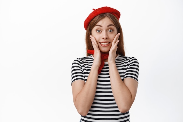 Free Photo portrait of amused girl in french beret, holding hands on face, smiling and looking with excitement at special promo offer, reacting to big announcement, white wall