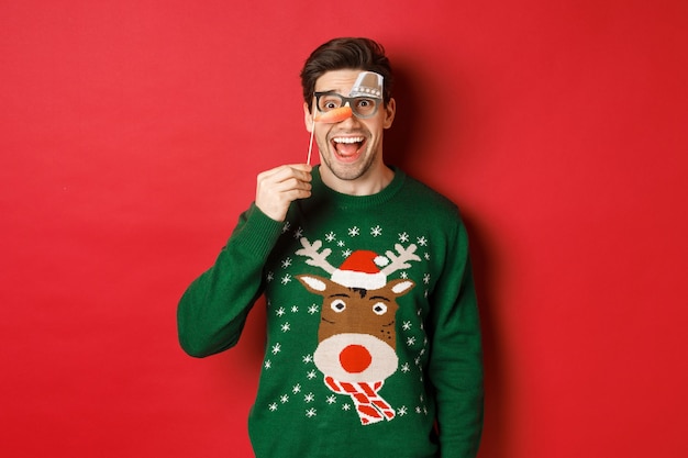 Portrait of amused handsome man in christmas sweater, holding funny party mask and smiling, celebrating winter holidays, standing over red background