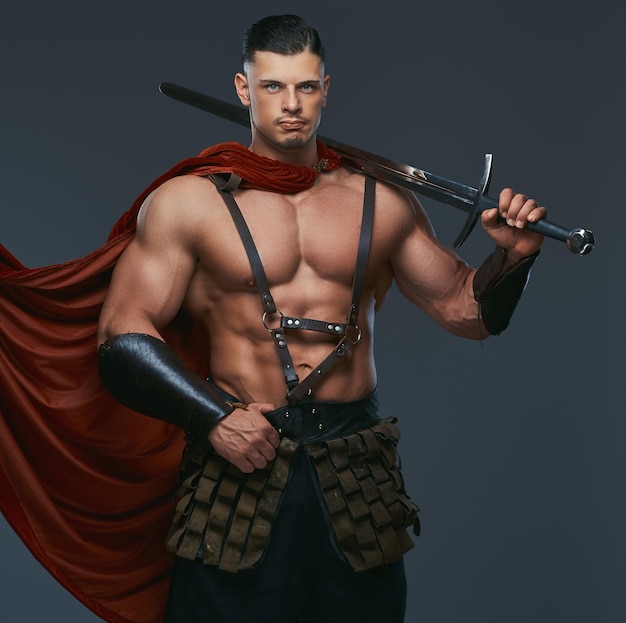 Portrait of ancient Greece warrior with a muscular body dressed in battle uniforms holds sword on his shoulder. Isolated on a gray background.