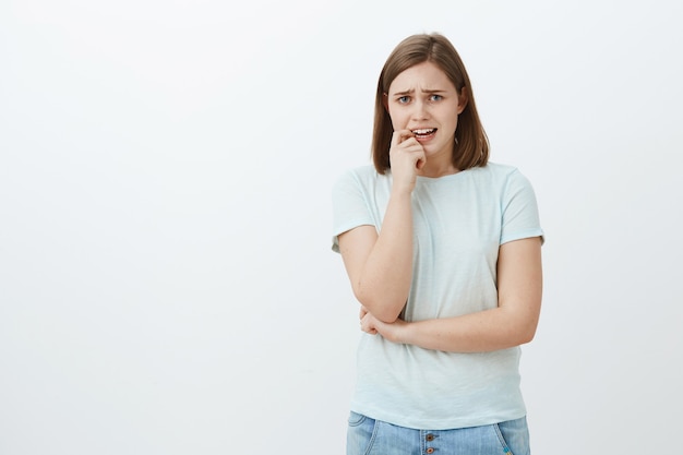 Portrait of anxious timid woman in casual outfit frowning whining and biting fingernail from nervous feelings standing insecure and worried over grey wall