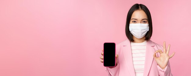 Portrait of asian businesswoman in medical face mask covid19 showing smartphone screen and okay sign pink background