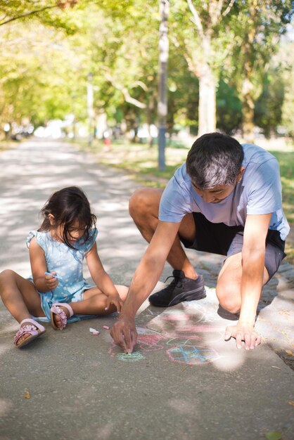 Portrait of Asian dad developing kids drawing skills. Happy man and little girl sitting on pavement in public park and drawing together with colorful crayons. Leisure in summer and parenthood concept