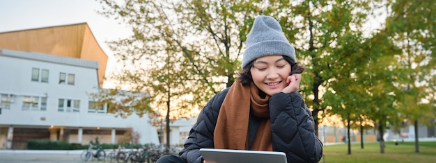 Free Photo portrait of asian girl relaxing in park watching videos or reading on digital tablet sitting on