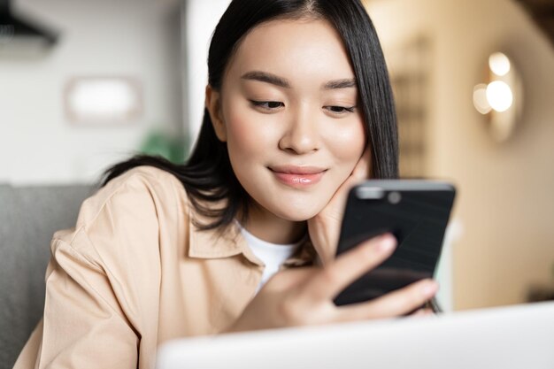 Portrait of asian girl sitting with laptop checking her phone and smiling browsing websites on smart...