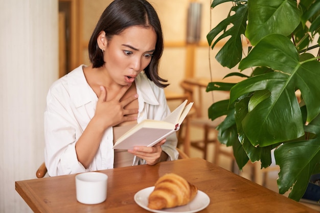 Free photo portrait of asian woman looking shocked at book pages reading something interesting concentrating si