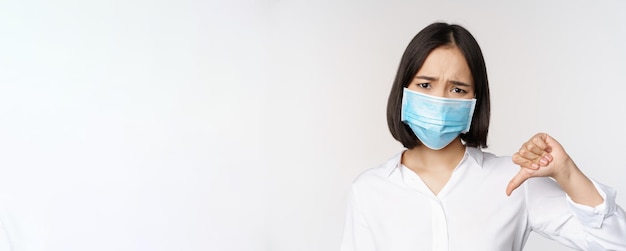 Portrait of asian woman in medical face mask showing thumbs down with disappointed tired face expres