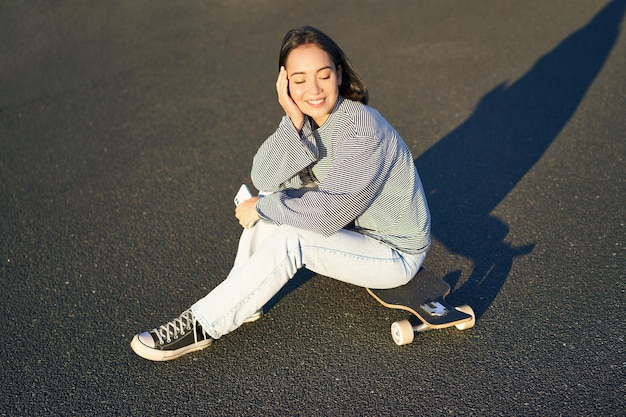 Free photo portrait of asian woman sitting on skateboard skating on her cruiser longboard using smartphone app