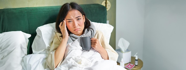 Free photo portrait of asian woman with headache catching col staying on sick leave at home lying in bed