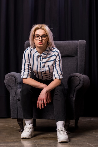 Portrait of an attractive blonde young woman sitting on armchair looking at camera