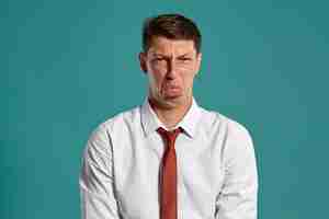 Free photo portrait of an attractive brunet male with brown eyes, wearing in a classic white shirt and red tie. he is acting like does not like something while posing in a studio against a blue background. conce