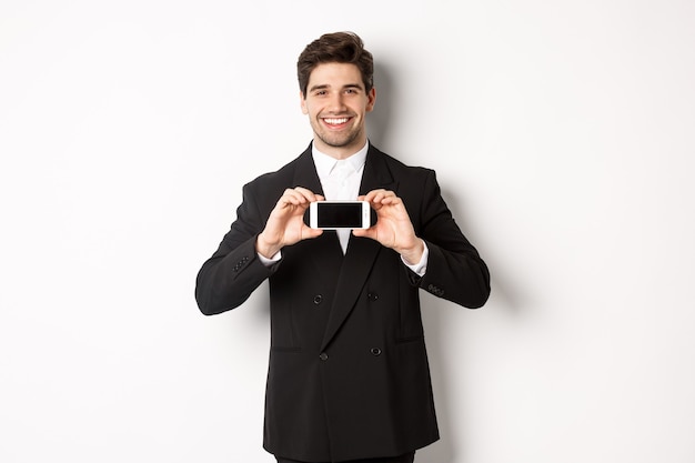 Free photo portrait of attractive businessman in black suit, holding smartphone horizontally and showing screen, smiling pleased, standing against white background.