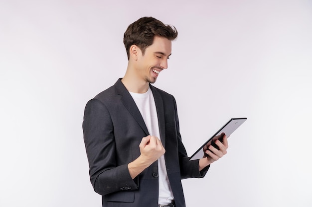 Portrait of attractive cheerful businessman doing winner gesture clenching fist and using device app searching web isolated over white color background