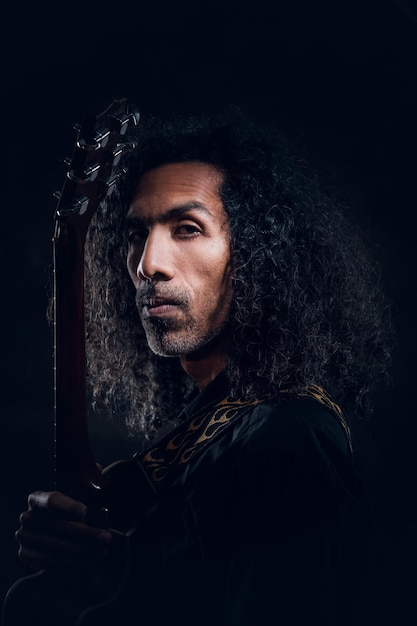 Portrait of attractive curly man with guitar at dark photo studio.