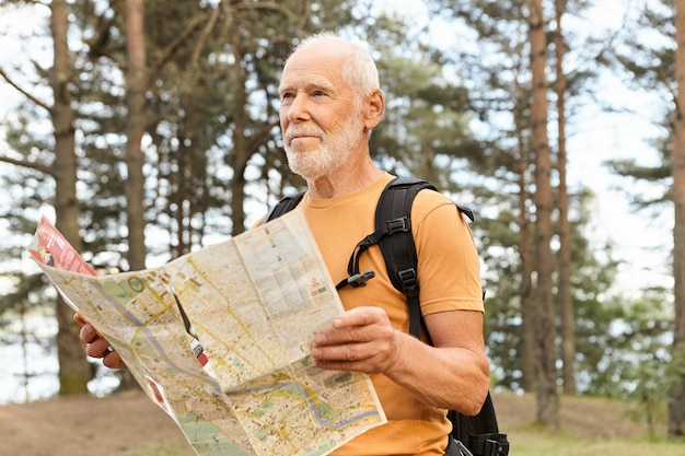 Free Photo portrait of attractive elderly male traveler backpacking using map, searching for right route. bearded senior caucasian male carrying rucksack thinking where to go, being at crossroads in forest