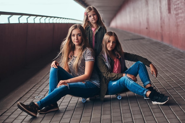 Free photo portrait of an attractive family. mother and her daughters sitting together on a skateboard at a bridge footway.