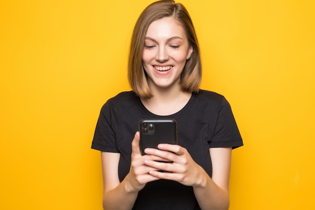 Portrait of attractive girl holding smart phone in hands, checking email, using 5G internet, typing sms, standing over yellow wall