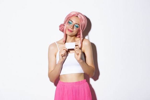 Portrait of attractive girl in pink wig, dreaming about shopping, holding credit card and smiling pleased, standing.