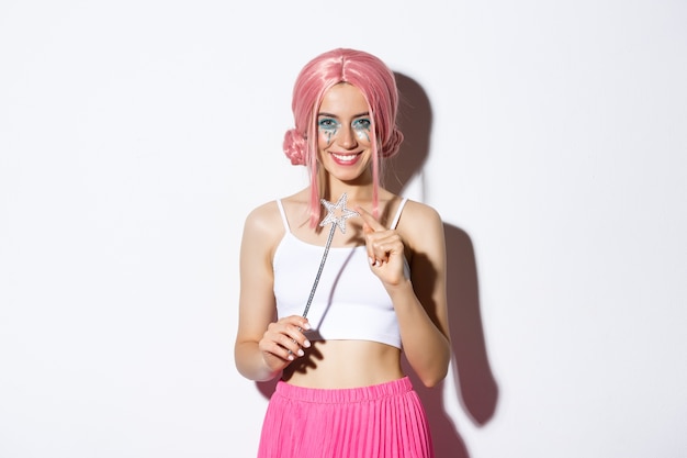 Free photo portrait of attractive girl with pink wig and bright makeup, dressed up as a fairy for halloween party, holding magic wand and smiling.