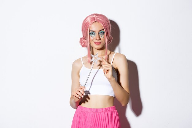 Portrait of attractive girl with pink wig and bright makeup, dressed up as a fairy for halloween party, holding magic wand and smiling