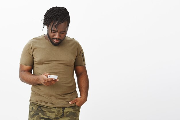 Portrait of attractive guy in a brown t-shirt posing against the white wall with his phone