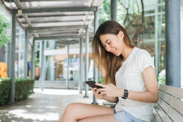 Portrait of attractive happy asian woman holding smartphone while sitting on roadside at the city 