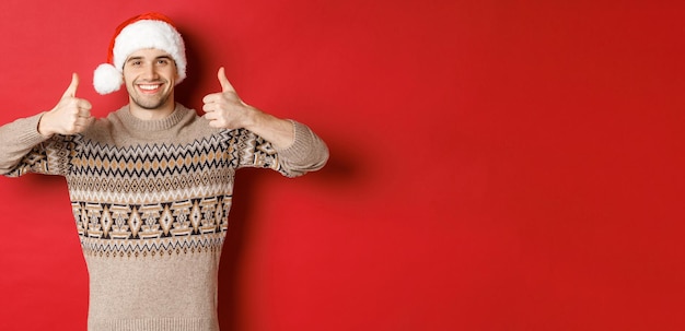 Free Photo portrait of attractive happy man in santa hat and sweater showing thumbsup in approval and smiling wishing merry christmas standing over red background