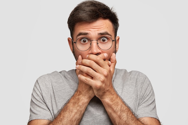 Free photo portrait of attractive male with frightened expression, covers mouth with both palms, being in stupor, notices something awful in front, dressed in grey clothes, isolated over white wall