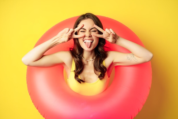 Free photo portrait of attractive modern girl inside trendy pink beach swimming ring, showing peace v-sign and smiling happy, standing over yellow background