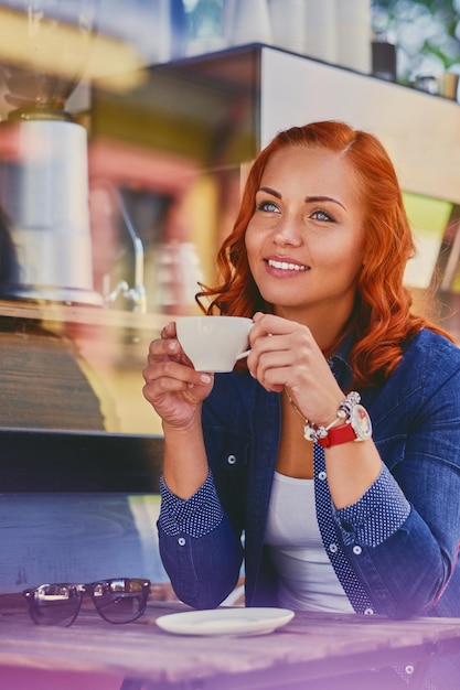 Free photo portrait of attractive redhead female drinks coffee in a cafe.