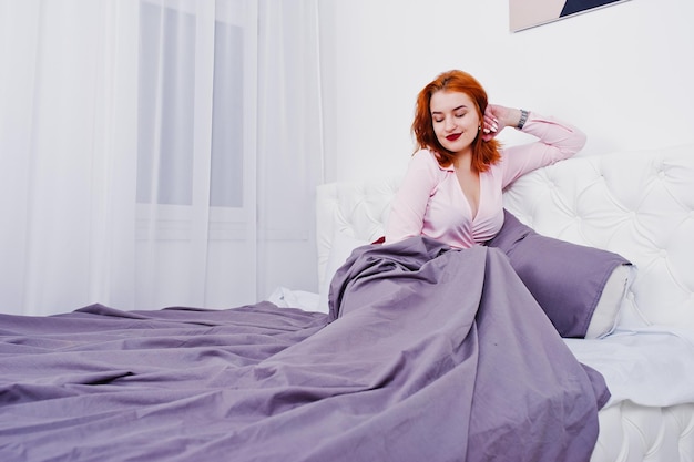 Portrait of an attractive redheaded girl laying on the bed and covering herself with a purple blanket