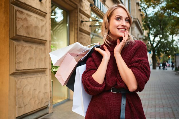 Portrait of attractive smiling blond girl with shopping bags happily looking away on city street