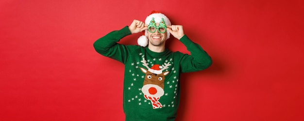 Portrait of attractive smiling man in santa hat party glasses and sweater celebrating new year holid...