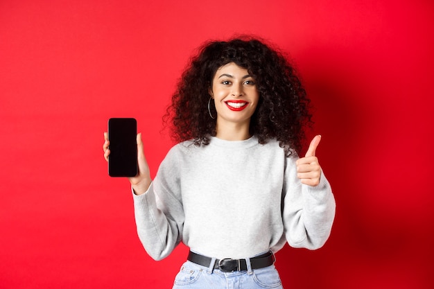 Free photo portrait of attractive smiling woman with curly hair, showing empty mobile phone screen and thumb-up, recommending online promo, standing on red background.