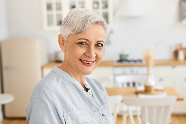 Free photo portrait of attractive stylish senior caucasian female pensioner with pixie short hairstyle spending day at home, standing in living room wearing elegant blue dress, smiling happily