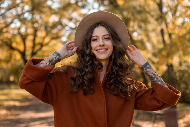 Portrait of attractive stylish smiling woman with long curly hair walking in park dressed in warm brown coat autumn trendy fashion, street style wearing hat