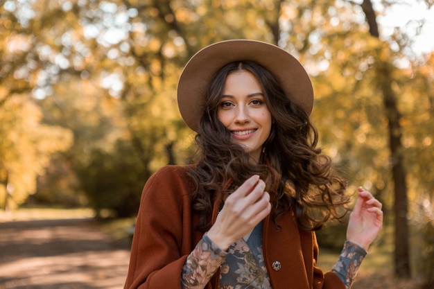 Portrait of attractive stylish smiling woman with long curly hair walking in park dressed in warm brown coat autumn trendy fashion, street style wearing hat