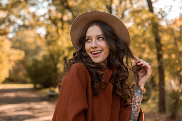 Free photo portrait of attractive stylish smiling woman with long curly hair walking in park dressed in warm brown coat autumn trendy fashion, street style wearing hat