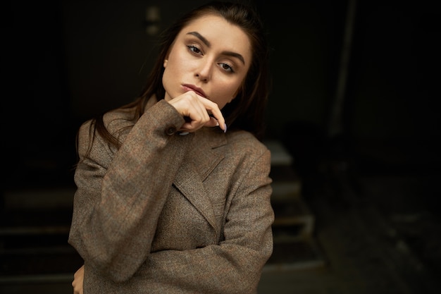 Portrait of attractive stylish young businesswoman having confident serious look staring at camera, holding hand on her chin. Beautiful thoughtful female posing isolated in warm oversize jacket