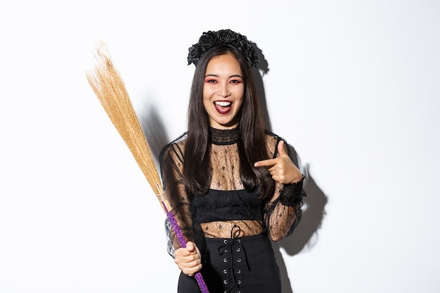 Free Photo portrait of attractive woman in witch costume celebrating halloween, pointing at broom, standing over white background.