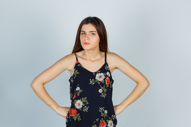 Portrait of attractive young lady holding hands on waist in blouse and looking serious front view