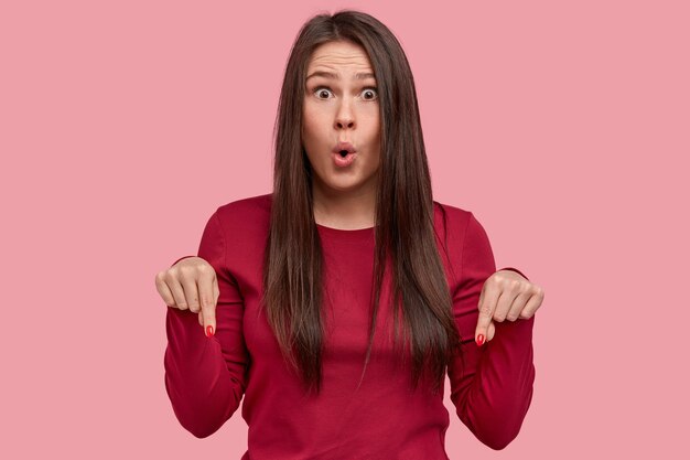 Portrait of attractive young woman has surprised face and opened mouth, points down at floor, wears casual clothes