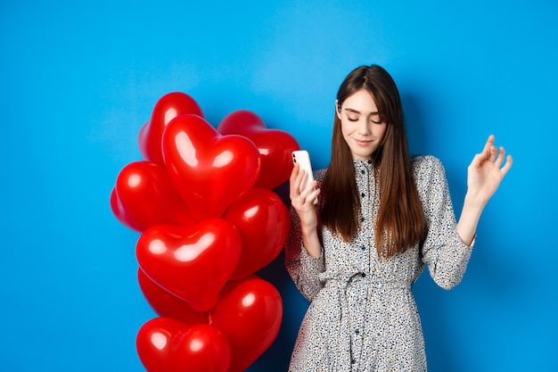 Free photo portrait of attractive young woman holding smartphone and dancing near valentines red balloons, standing on blue background