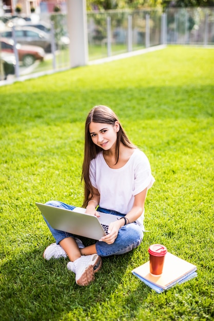 Portrait of attractive young woman sitting on green grass in park with legs crossed during summer day while using laptop