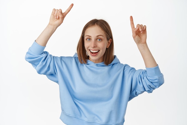 Free photo portrait of attractive young woman smiling, pointing fingers at logo, showing sale discount banner, advertisement up, standing in blue hoodie, recommend click on link, white background.