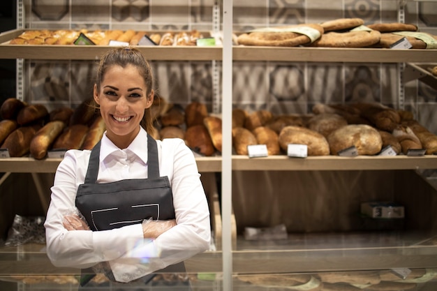 Free photo portrait of bakery seller with arms crossed standing in front of shelf full of bred bagels and pastry