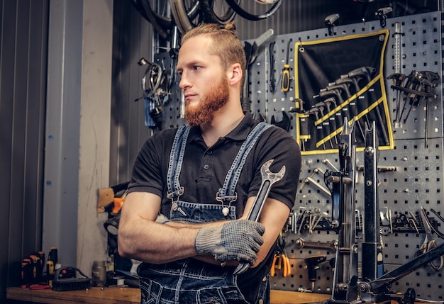 Portrait of bearded bicycle mechanic with crossed arms holds cup key over tool stand background in a workshop.