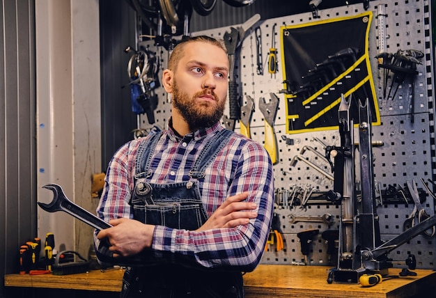 Portrait of bearded bicycle mechanic with crossed arms holds cup key over tool stand background in a workshop.