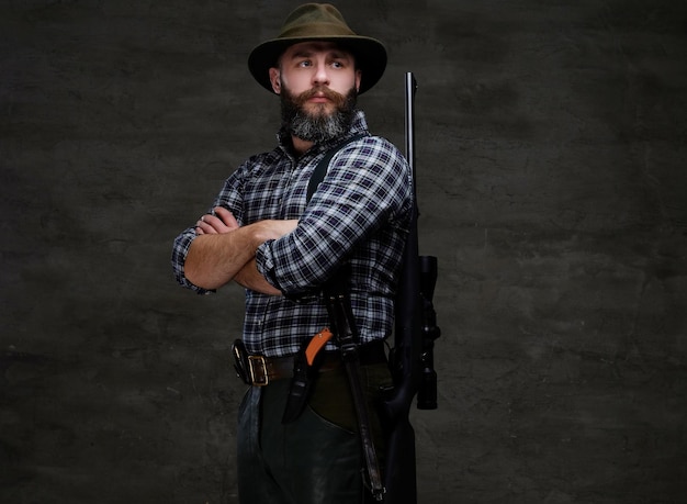 Free photo portrait of a bearded hunter in a fleece shirt and hat standing with a rifle behind his back. isolated on a dark background.