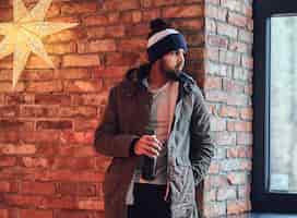 Free photo portrait of bearded indian hipster male dressed in a warm jacket and a hat, drinks coffee from a red cup near the wall of a red brick.